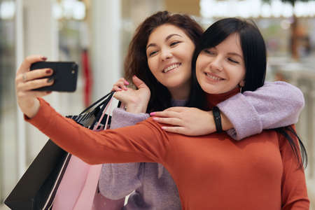 Beautiful girls with shopping bags taking selfie in shopping mall, looking at smart phone screen...