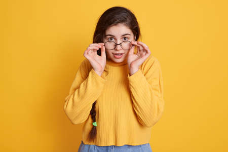 Attractive woman with new glasses keeping hands on her frame, posing isolated over yellow background...