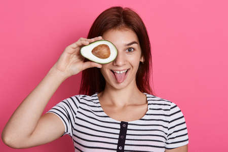 Happy young woman holding half of avocado, covering her eye with healthy fruit, showing her tongue, wearing striped t shirt, posing isolated over pink background.の写真素材