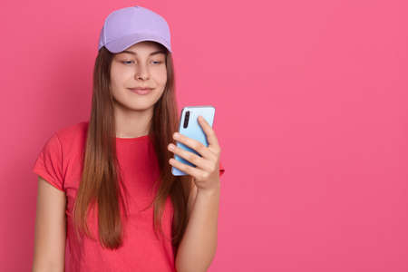 Smart female with mobile phone in hand, expressing positive emotions, standing smiling over pink...