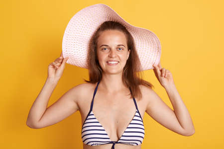Portrait of smiling dark haired woman woman wearing straw hat and striped white and black swimming suit, looking at camera with charming smile isolated over yellow background.の写真素材
