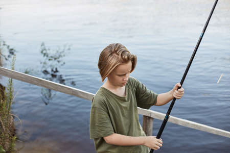 Happy boy fishing from wooden placing, holding fishing rod in hands and being ready to cast line,...