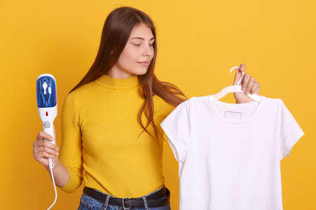 Attractive young Caucasian woman steaming clothes against yellow background, holding white t shirt on hangers, looking at her outfit, being ready to iron attire.の写真素材