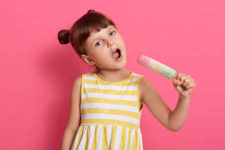 Pretty baby girl kid holding big ice cream as microphone and singing, female child imagines she singer and sings with water ice in hands against pink wall.の写真素材