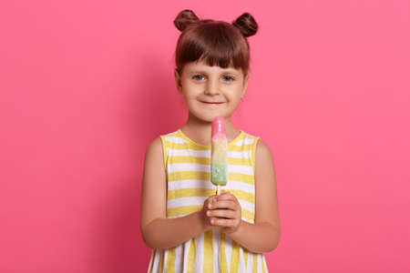Little girl with big fruit ice cream standing against rosy wall and looking directly at camera, wearing summer outfit, having two knots, expressing positive emotions.の写真素材