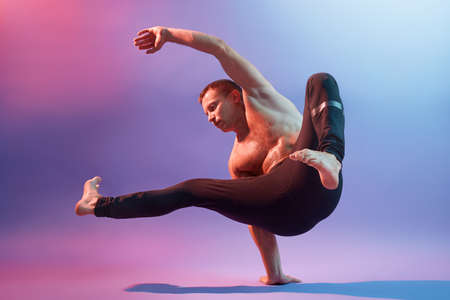 Young sportsman acrobat or gymnast posing against neon wall, showing his power, stands on one hand,...