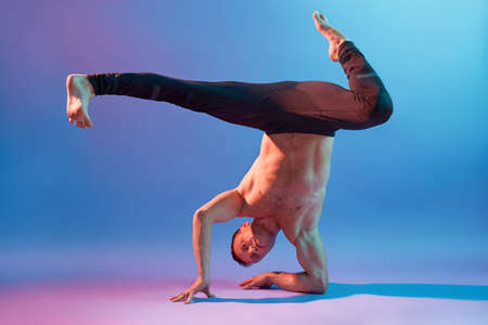 Male yoga standing on hands upside down, wearing black trousers, posing topless isolated over neon...