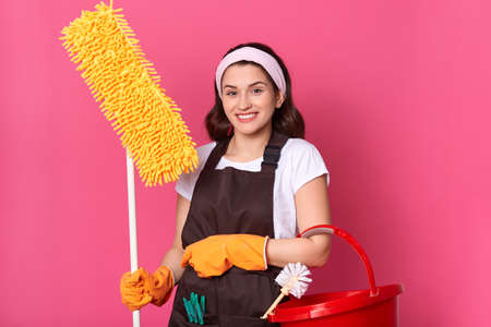 Front view of smiling young woman housewife in casual clothes and apron, doing housework isolated on pink background, holding yellow mop and red bucket.の写真素材