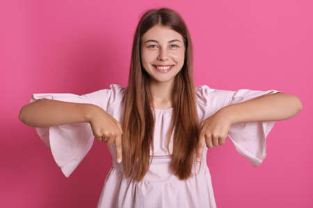 Charming carefree woman with positive expression pointing down with both index fingers, wearing beautiful dress, has broad smile, isolated over pink background.の写真素材