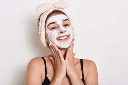 Smiling woman with hairband on her head applied mask to skin of her face, looking smiling directly...