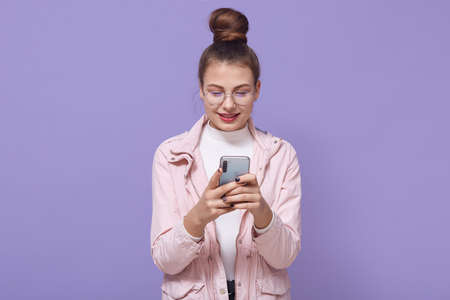 Young pretty European female isolated over lilac background standing in pale pink jacket, texting on...