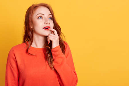 Beautiful trendy girl in orange sweater being deep in thoughts, looking in distance with finger on...