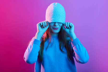 Attractive woman pulling hat over her eyes wearing casual sweater posing isolated over pink neon background, young lady with long hair having fun,の写真素材
