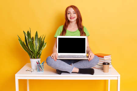 Beautiful young red haired college student sitting on white table with lap top near flower, dressed...