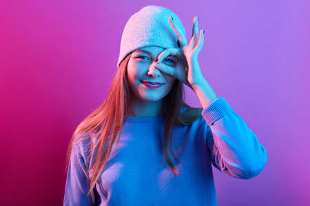 Portrait of excited young woman dressed casual sweater and knitted hat, showing ok sign against her...