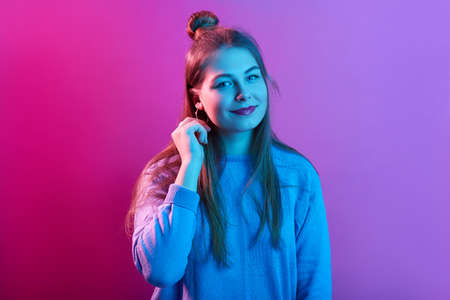 Pleasant looking young woman wears casual sweater, has hair bun, looks with calm facial expressions, poses against pink neon background, lady keeps hand near ear.の写真素材