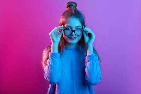 Adorable female touching frames of glasses, smiling delighted and looking at camera against pink neon wall, girl with hair bun and wearing casual sweater.の写真素材