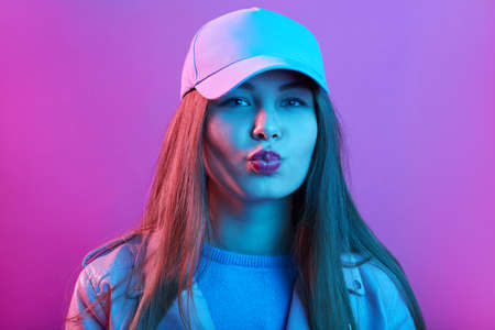 Fashion model girl isolated over pink neon background, beauty stylish woman with long hair posing with rounded lips, making kiss gesture, wearing baseball cap.の写真素材