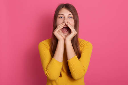 Cheerful funny young woman wearing yellow sweater screaming with hand gesture near mouth isolated...