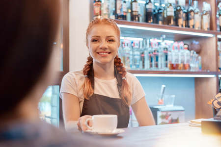 Happy young barista offering cup of coffee while smiling at camera or to her client, wearing white casual t shirt and brown apron, posing at counter.の写真素材