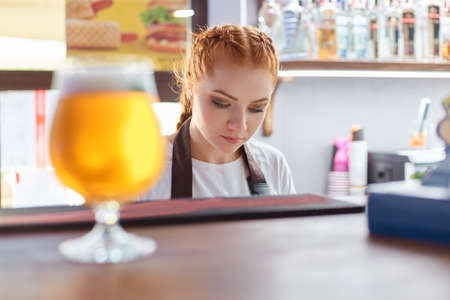 Attractive bartender selling beer in bar, red haired barkeeper, glass of beer on dark counter, young female barman working in pub, wearing white casual t shirt.の写真素材