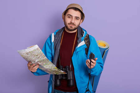 Bearded young explorer posing against lilac wall with compass and map, has binocular on his neck,...