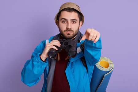 Portrait of serious young European tourist guy with bag and binoculars isolated over lilac background, looking at camera and pointing with index finger.の写真素材