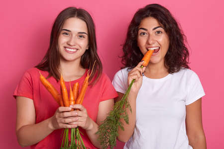 Picture of cheerful positive beautiful friends standing isolated over pink background, opening mouth, holding carrots in hands, wearing casual attires.の写真素材