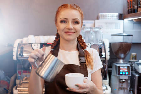 Waitress taking order from her customer in cafe, red haired barista wearing white t shirt and brown apron, looking smiling directly at camera, holding turk and ceramic cup.の写真素材