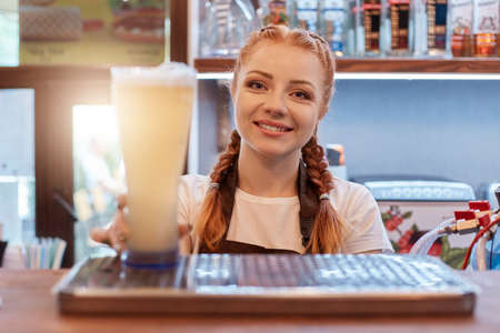 Red haired positive woman with pigtails holding beer glass while working at bar, attractive bartender wearing white t shirt and apron, stands with showcase with booze.の写真素材