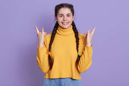 Young brunette woman posing isolated over purple background, showing rock gesture with fingers and...