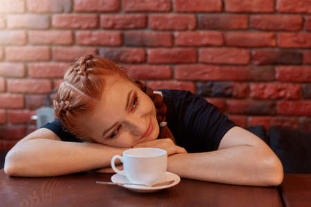 Beautiful woman with cup of coffee lies on table and looking smiling aside, tired student has break, posing against brick wall in black wall, red haired lady resting in cafeteria.の写真素材