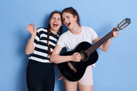 Young band rehearsing, girls in casual attires singing songs loudly, standing against blue wall,...