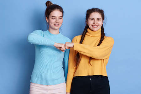 Two caucasian women fist bumping against blue wall, two friends greeting, females wearing casual shirts, have dark hair, expressing happiness and positive emotions.の写真素材