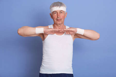 Senior man stretching hands indoors against blue background, warming up before training or playing...