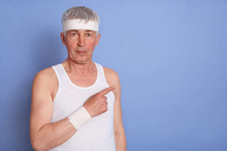 Senior man in white t shirt looking at camera, pointing aside with his forefinger, copy space for...