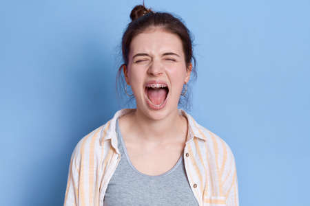 Young emotional angry woman screaming isolated over blue background, keeping eyes closed, looks sad, wearing white shirt and gray t-shirt, brunette lady with knot yelling angrily.の写真素材