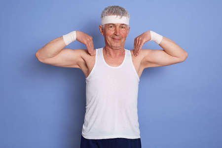 Indoor shot of happy energetic senior man enjoying physical training against blue wall, doing physical exercise, holding fingers on his shoulders, looking at camera with smile.の写真素材