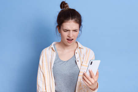 Winsome young brunette woman feeling doubtful and confused, wondering which decision to make, using smart phone, keeping mouth opened, posing isolated over blue background.の写真素材