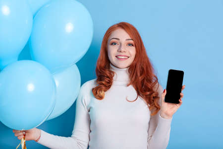 Smiling pretty young woman in white shirt, holds blue balloons and mobile phone with blank empty...