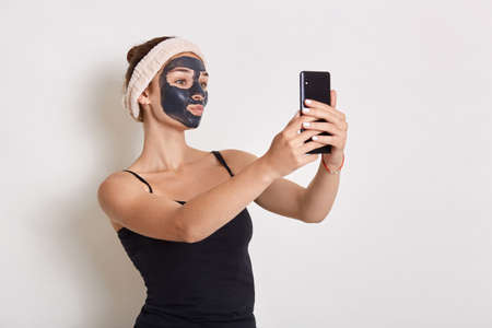 Portrait of girl with black cosmetic mask on her face holding modern smart phone in hand and looking...