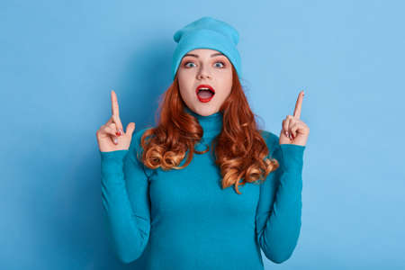Teen girl pointing two fingers up at empty copy space, posing isolated over blue background, red...