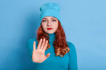 Confident redhead woman keeps palms towards camera, has displeased expression, looks at camera,...