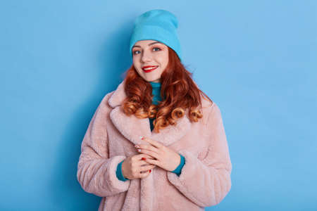 Beautiful woman wears pink fur coat and blue cap, smiles toothily, wears red lipstick, looks at...