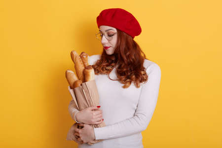 Charming woman with red hair, wears beret and white sweater, looks at long loaf in her head, isolated over yellow background, copy space for promo, romantic female with long bread baguettes.の写真素材