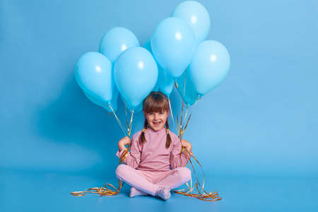 Cute little girl sitting on floor with bunches of helium balloons, looks at camera and laughing, wearing casual attire, being in good mood, sitting on floor with crossed legs against blue wall.の写真素材