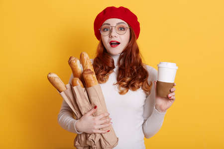 Portrait of astonished woman wearing beret, white shirt and glasses, holding paper bags with fresh baguettes and coffee to go, female with opened mouth and surprised facial expression.の写真素材