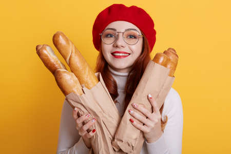 Portrait of cheerful woman wearing beret, white shirt and spectacles, looking smiling at camera, holding paper bags with fresh baguettes, showing toothy smile, expressing positive emotions.の写真素材