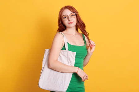 Young red haired girl with white blank cotton shopper tote bag wearing green t-shirt, mock-up of blank bag, standing against yellow wall, lady with red hair in spectacles.の写真素材