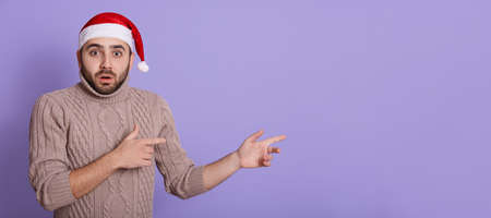 Serious and shocked young Santa man in beige sweater and Christmas hat, pointing index fingers aside...